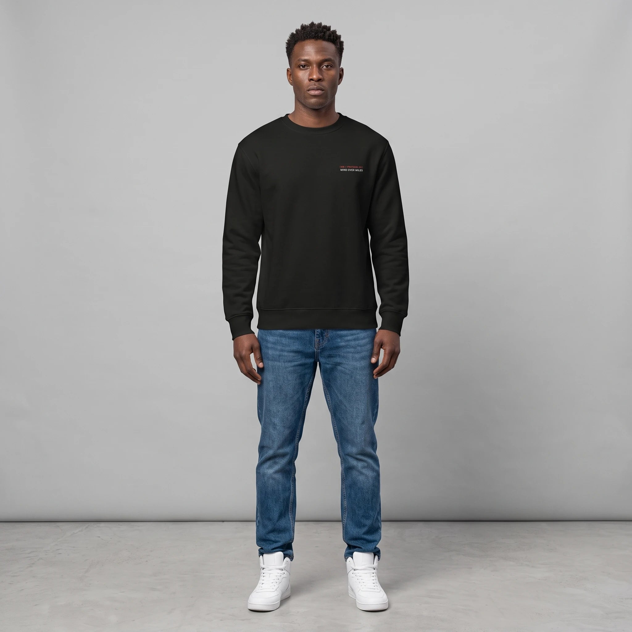 Full-length front view of a male model with short dark hair standing against a grey background in a studio setting, wearing a black crewneck sweatshirt with small chest text, paired with blue jeans and white sneakers.