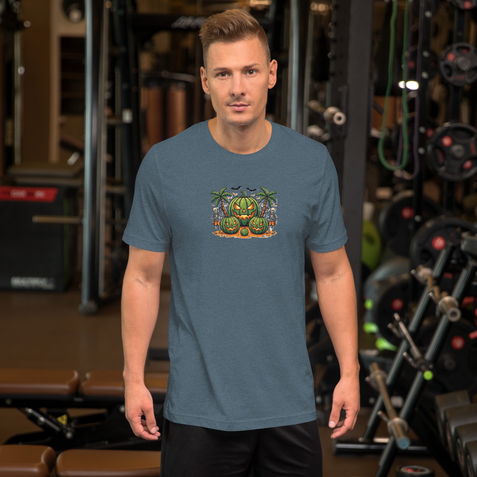 Front view of a male model standing in a gym, wearing a heather blue men's Summerween graphic t-shirt with a tropical watermelon jack-o'-lantern and skeletons design.