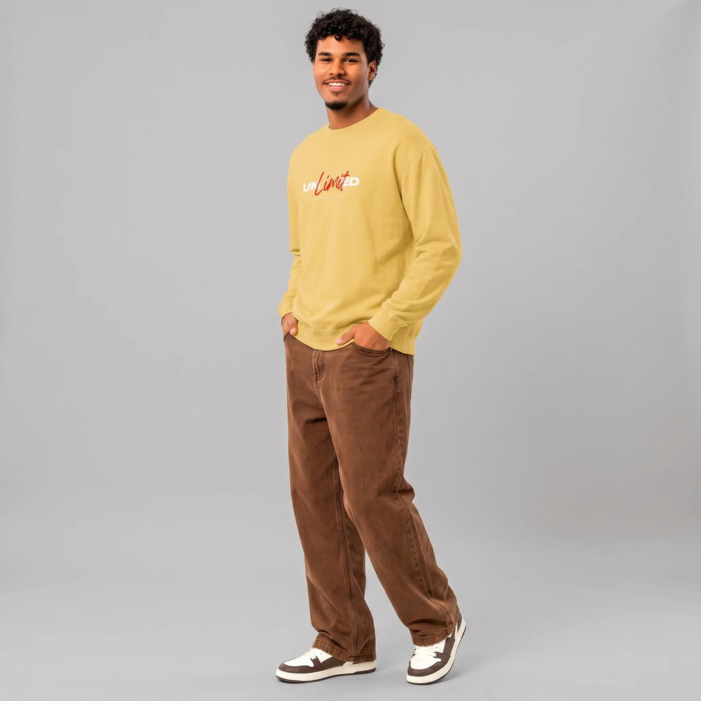 Full-length front view of a smiling male model with curly hair against a grey background, wearing a yellow crewneck sweatshirt with 'UNLIMITED' printed across the chest in red script and white block letters, paired with loose-fitting brown pants and sneakers.