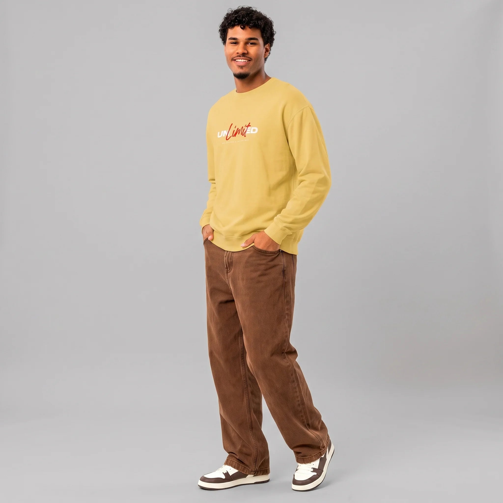 Full-length front view of a smiling male model with curly hair against a grey background, wearing a yellow crewneck sweatshirt with 'UNLIMITED' printed across the chest in red script and white block letters, paired with loose-fitting brown pants and sneakers.