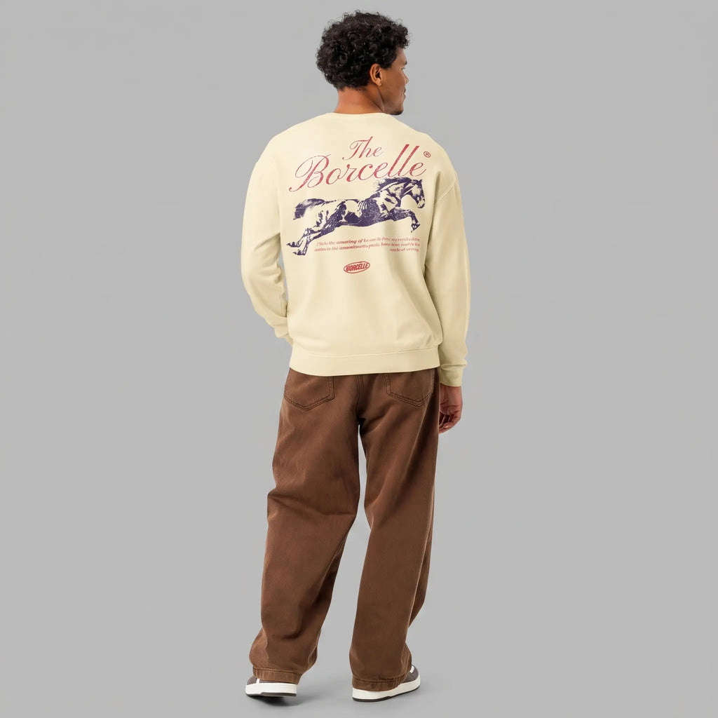 Back view of a male model with dark curly hair, standing against a grey studio background. He is wearing an ivory/cream-colored crewneck sweatshirt and loose-fitting brown pants. The back of the sweatshirt features a large, vintage-style graphic. At the top, in large red cursive script, is the text 'The Borcelle'. Below this is a detailed, dark (purple) silhouette of a leaping or galloping horse. Under the horse, small red text reads: 'From the pounding of horses' hooves to powerful leaps, rodeo is the embo