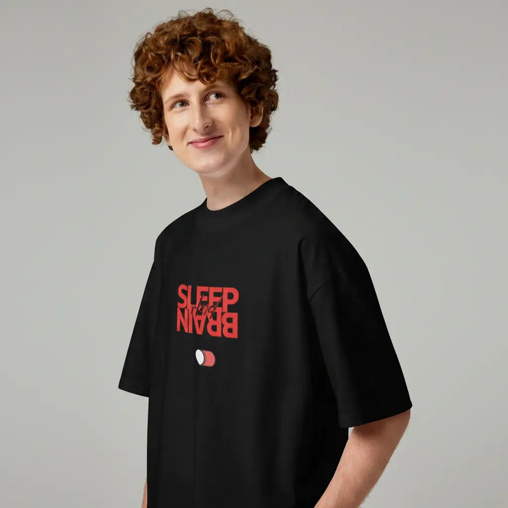 Side angle view of a smiling man wearing an oversized black organic cotton t-shirt. The shirt clearly shows the red Sleep and Brain toggle switch graphic on the chest.
