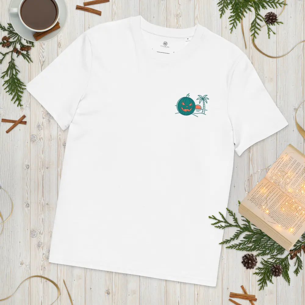 Flat lay mockup of a white organic cotton unisex t-shirt featuring a Summerween tropical green jack-o-lantern and palm tree graphic on the left chest, styled on a wooden surface with an open book, coffee cup, and pine branches.