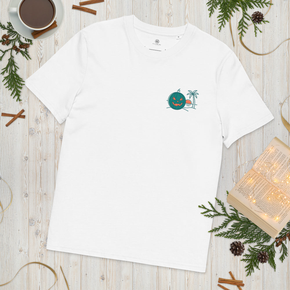 Flat lay mockup of a white organic cotton unisex t-shirt featuring a Summerween tropical green jack-o-lantern and palm tree graphic on the left chest, styled on a wooden surface with an open book, coffee cup, and pine branches.