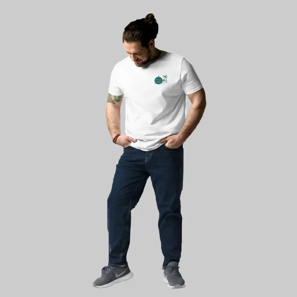 Full-body shot of a male model wearing a white organic cotton unisex t-shirt featuring a Summerween tropical green jack-o-lantern and palm tree graphic on the left chest, paired with blue jeans.