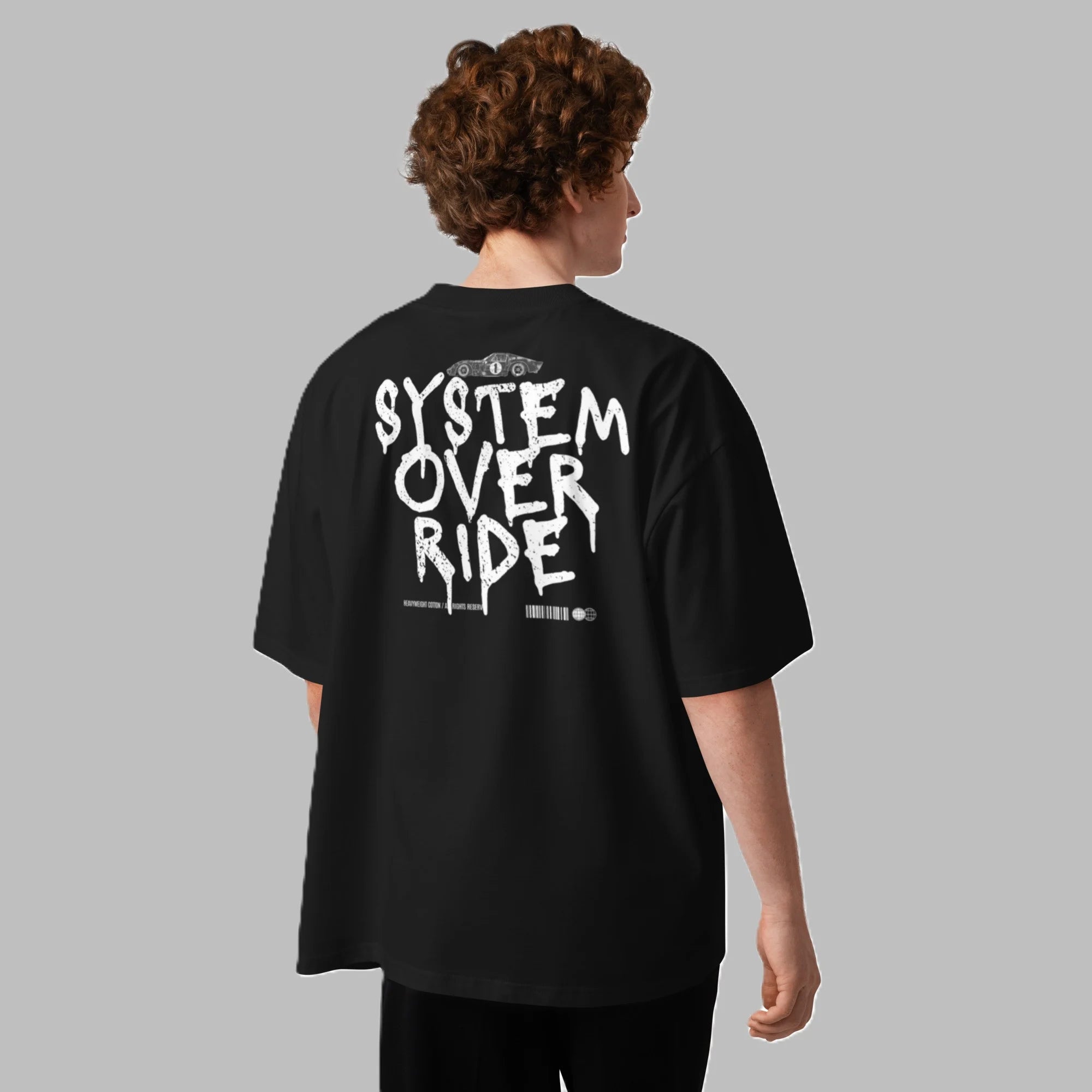 A male model from behind, wearing a black oversized organic cotton streetwear t-shirt featuring a large white 'SYSTEM OVERRIDE' graphic print across the back.