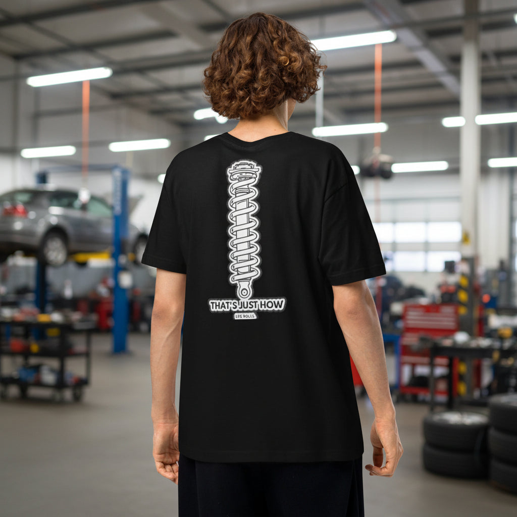 Back view of a male model in an auto repair garage wearing a black car enthusiast t-shirt featuring a large white coilover suspension graphic and the text That's Just How Life Rolls.